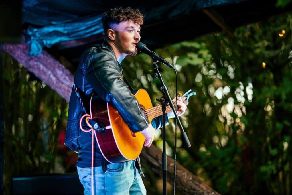 The Falcon Inn Musician playing acoustic guitar on stage, wearing a leather jacket.